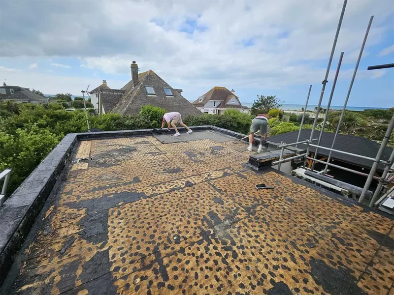 Two of the Trinity Roofing Sussex team stripping an old flat roof back to the deck, with the Sussex coastline and the sea behind them