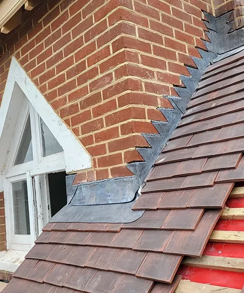 Stepped lead flashing along a roof verge beside a dormer window, red clay tiles surrounding