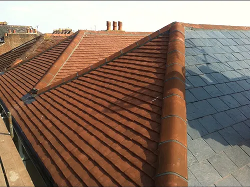 Hip roll lead detail on a red clay tile roof corner with chimney pots against blue sky