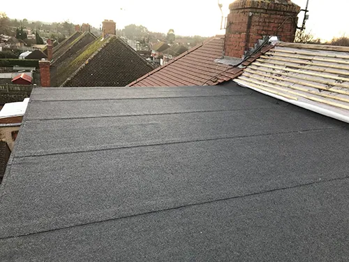 Worn clay tile ridge showing ageing and exposed felt during a repair assessment, chimney stacks beyond